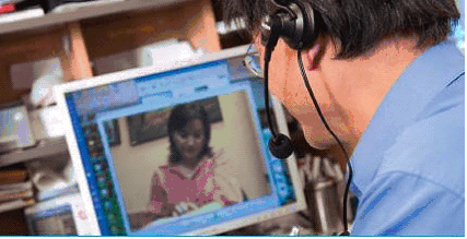 Man with a head set on looking at an image of a person on his computer.